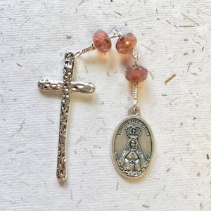 May include: A silver rosary with a silver cross and a silver pendant featuring the Virgin of Guadalupe. The beads are made of small, clear, iridescent glass beads in shades of pink and purple.