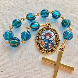 May include: A blue glass rosary with a gold-toned cross and a pendant featuring a stained glass image of Saint Peter. The pendant has the text "St. Peter" on it.