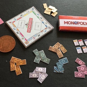 May include: A miniature Monopoly game set with a board, box, money, and Chance and Community Chest cards. The board is white with a blue and red border. The box is red with the word "MONOPOLY" in white letters. The money is in various denominations, including $1, $5, $10, $20, $50, $100, and $500. The Chance and Community Chest cards are small and rectangular.
