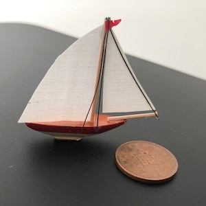 Puede incluir: Un pequeño velero de madera con una vela blanca y una bandera roja. El velero es del tamaño de una moneda de un euro.