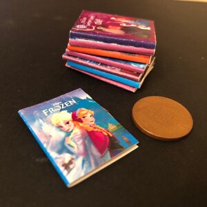 以下が含まれることがあります： カラフルな表紙のミニチュアペーパーバックの積み重ね。中には、エルサとアナが登場するアナと雪の女王をテーマにした本も含まれています。画像には銅色のコインも写っています。