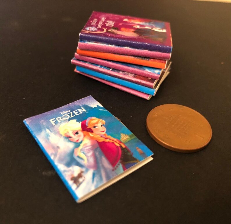 以下が含まれることがあります： カラフルな表紙のミニチュアペーパーバックの積み重ね。中には、エルサとアナが登場するアナと雪の女王をテーマにした本も含まれています。画像には銅色のコインも写っています。