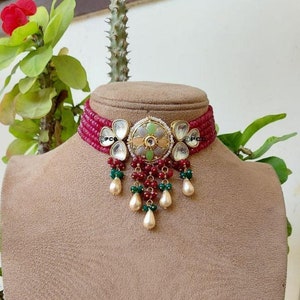 May include: A red and green beaded choker necklace with a gold and white floral pendant. The pendant has a round center with a smaller round stone in the center. The necklace has a dangling design with pearls and green beads.