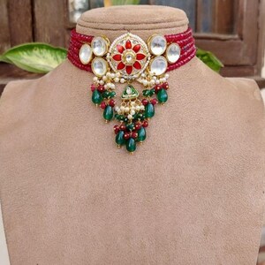 May include: A maroon beaded choker necklace with a central floral pendant featuring red and white accents, surrounded by clear stones. Matching earrings with similar designs complete the set. The jewelry is displayed on a mannequin.