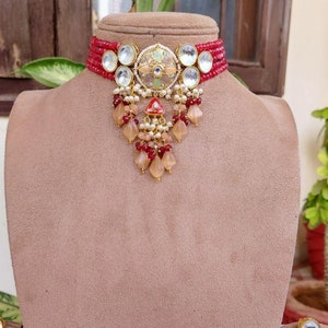 May include: A red beaded choker necklace and matching earrings set. The necklace features a central pendant with a floral design, clear stones, and dangling beads. The earrings mirror the pendant design, with a circular shape and dangling beads.