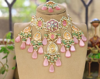 May include: A pink and green Indian bridal necklace set with a gold base and intricate detailing. The necklace features a large central pendant with a floral design and is adorned with pearls and gemstones. The earrings are similarly designed with a smaller pendant and matching embellishments.