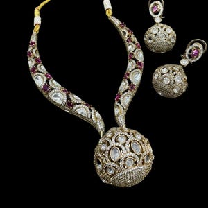 May include: A gold-toned jewelry set featuring a necklace and matching earrings. The necklace has a large, round pendant and a curved design with clear and red gemstones. The earrings are round with similar gemstone detailing. The set is displayed against a black background.