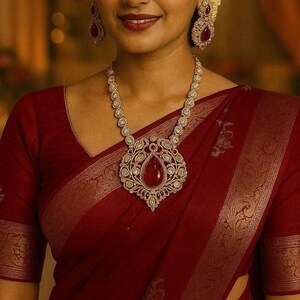 May include: A woman wearing a maroon sari and ornate jewelry. The necklace features a large, teardrop-shaped red gemstone pendant surrounded by a detailed, gold-toned setting. Matching earrings complete the set. The sari has gold-colored trim.