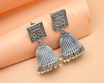 May include: A pair of silver earrings with a square design and a bell-shaped dangle. The earrings have a floral pattern and are decorated with small white beads.