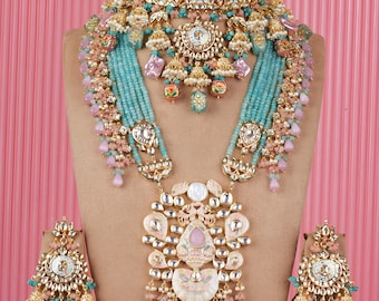 May include: A detailed gold-coloured jewellery set comprising a choker, long necklace, and matching earrings. The set incorporates turquoise, pink, and white accents, with intricate detailing and beadwork. The jewellery is displayed on a neutral-toned bust against a pink backdrop.
