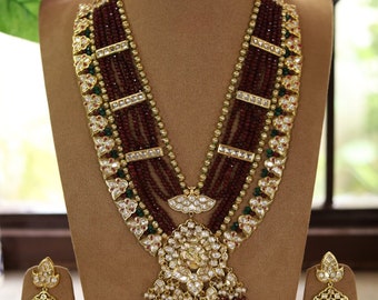 May include: A gold necklace set with a large pendant featuring a floral design and red and green beads. The necklace is on a brown display stand. The earrings are also gold with a floral design and red and green beads.