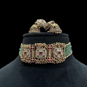 May include: A gold-toned choker necklace and matching earrings set. The choker features square and rectangular jewel-encrusted elements with red and clear stones, and green beaded accents. The earrings have a similar design. The jewelry is displayed on a black mannequin head.