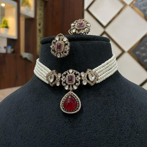 Może przedstawiać: Trzyczęściowy zestaw biżuterii z naszyjnikiem z pereł z centralną zawieszką oraz pasującymi kolczykami i pierścionkiem. Zestaw ma metal w kolorze złotym z czerwonymi i przezroczystymi kamieniami.