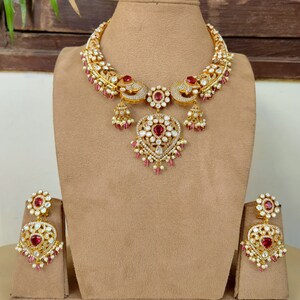 May include: A gold necklace with a peacock design and a heart-shaped pendant. The necklace is adorned with white and pink stones. Matching earrings with a similar design are also included.