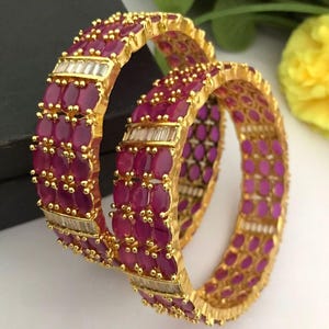 Op de afbeelding: Twee goudkleurige armbanden met robijnrode stenen en heldere rechthoekige accenten. De armbanden zijn op elkaar gestapeld, wat het gedetailleerde ontwerp en het contrast tussen het goud en de rode stenen laat zien. De sieraden staan tegen een neutrale achtergrond.