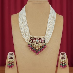 May include: A multi-strand white beaded necklace with a burgundy and gold pendant, and matching earrings. The pendant and earrings feature a floral design with red and green accents, and a cluster of red beads.