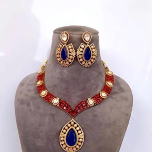 May include: A jewelry set consisting of a necklace and matching earrings. The necklace features a V-shaped band of red stones with white accents, ending in a large teardrop-shaped blue pendant. The earrings mirror the teardrop design.