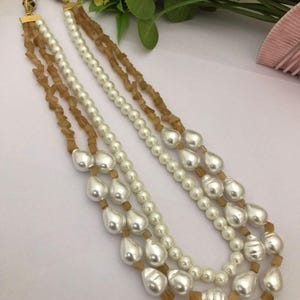 May include: A three-strand pearl necklace with brown and gold beads. The necklace features large, teardrop-shaped pearls.