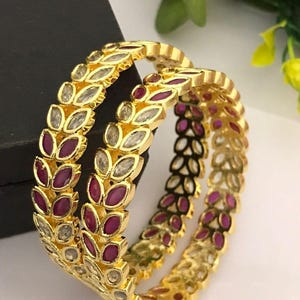 May include: Two gold bangles featuring a leaf design, each leaf set with clear and ruby-red gemstones. The bangles are presented on a black surface, highlighting their detailed craftsmanship and vivid colours. The gemstones are set in a repeating pattern.