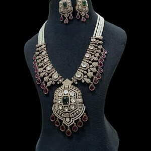 May include: A luxurious necklace and earring set displayed on a black mannequin. The necklace features a large pendant with a central green stone, surrounded by clear and red gemstones. The earrings mirror the design, creating a cohesive jewelry set.