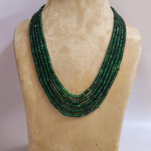 May include: A multi-strand necklace featuring small, faceted, emerald green beads. The necklace is draped on a mannequin, showcasing its layered design and rich color. The beads catch the light, creating a sparkling effect.