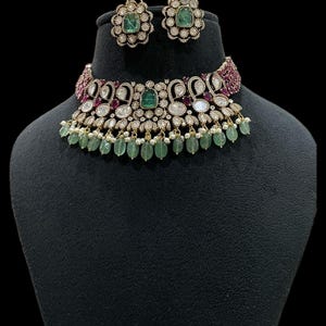 May include: A gold-toned necklace and matching earrings set. The choker-style necklace features emerald green and clear gemstones, with a fringe of green beads. The earrings have a floral design with a green gemstone center. The jewelry is displayed on a black mannequin.