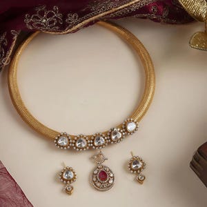 Puede incluir: Un conjunto de collar y pendientes dorados. El collar presenta una banda gruesa y tejida con una fila de piedras redondas y claras. El colgante tiene una piedra roja en el centro. Los pendientes tienen un diseño similar. Un tejido granate con ribete dorado está en el fondo.