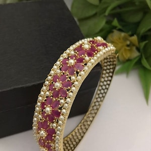 Può includere: Un braccialetto rigido in oro con un designo di fiori rosa e perle bianche. Il braccialetto è realizzato in metallo e ha un design delicato e intricato.