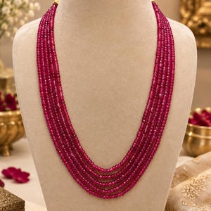 May include: A multi-strand necklace featuring five rows of small, faceted, deep magenta-red gemstone beads. The beads are uniform in size and shape, creating a layered, cascading effect as they drape down the center of the display form.