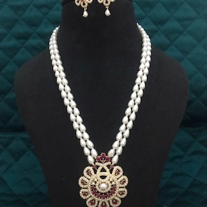 Può includere: Una collana di perle a due fili con un grande ciondolo e orecchini abbinati. Il ciondolo e gli orecchini presentano un design floreale color oro con pietre preziose rosse e trasparenti. La collana è esposta su un manichino nero.
