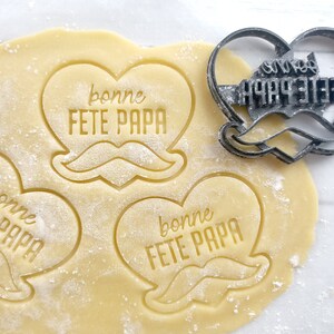 Peut inclure: Trois emportes-pièces en forme de cœur avec le texte "bonne fête papa" et un motif de moustache. Les emportes-pièces sont sur une surface de pâte blanche.