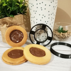 Puede incluir: Tres galletas redondas con relleno de chocolate, junto con un cortador de galletas negro y un anillo negro. Las galletas tienen un exterior marrón dorado y un centro cremoso marrón oscuro. Una taza blanca con un patrón de estrellas y una pequeña planta en un jarrón de vidrio están en el fondo.
