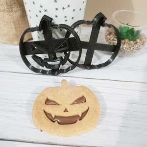Puede incluir: Dos cortadores de galletas de plástico negro en forma de calabaza con un diseño de cara de miedo. Una sola galleta en forma de calabaza con relleno de chocolate se muestra en primer plano.