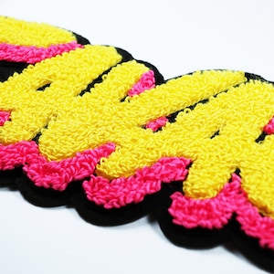 May include: A yellow and pink embroidered patch with the word "SWAG" in a bold, stylised font. The patch is outlined in black and has a fuzzy texture.