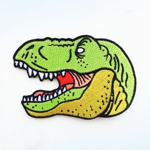 May include: Green and yellow embroidered iron-on patch of a dinosaur head with an open mouth and sharp teeth.