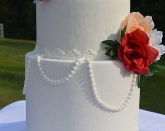 Puede incluir: Un pastel de boda blanco de tres niveles decorado con encaje blanco y detalles de perlas. El pastel está adornado con rosas rojas y melocotón y flores blancas.