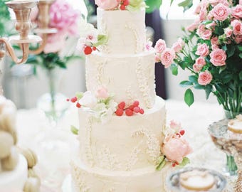 Puede incluir: Un pastel de boda blanco de cuatro pisos decorado con delicados detalles florales y rosas rosadas. El pastel está sobre un soporte para pastel blanco y rodeado de flores rosadas.