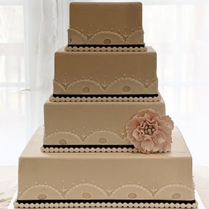 May include: A four-tiered wedding cake with white frosting and delicate lace-like designs. Each tier is separated by a black ribbon and a row of white dots. A single pink sugar flower sits on the top tier.