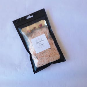 May include: A black, resealable bag filled with pink bath salts. The clear front of the bag reveals the product, with visible dried rose petals. A white label reads "Lavender Rose" and "Bath & Body Soak".