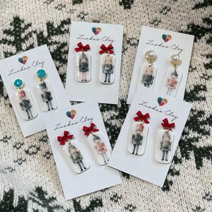 May include: A collection of handmade earrings featuring miniature soldier designs. Each pair is attached to a white card with a red bow and the text "Zuska's Clay". The earrings showcase various soldier uniforms and are set against a patterned background.