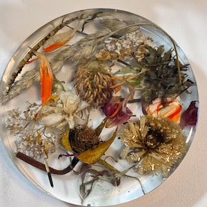 May include: A clear, round resin coaster with dried flowers and foliage embedded within. The botanical elements include orange, white, and brown dried flowers, and various grasses. The coaster is a decorative item for home decor.