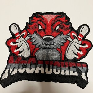 K&ouml;nnte beinhalten: Roter und schwarzer gestickter Aufn&auml;her mit einer Comicfigur, die zwei Bowling-Pins h&auml;lt. Die Figur hat einen roten K&ouml;rper, einen grauen Bart und einen schwarzen Umriss. Der Text "McCauley" ist in wei&szlig;en Buchstaben auf einem schwarzen und grauen Hintergrund geschrieben.