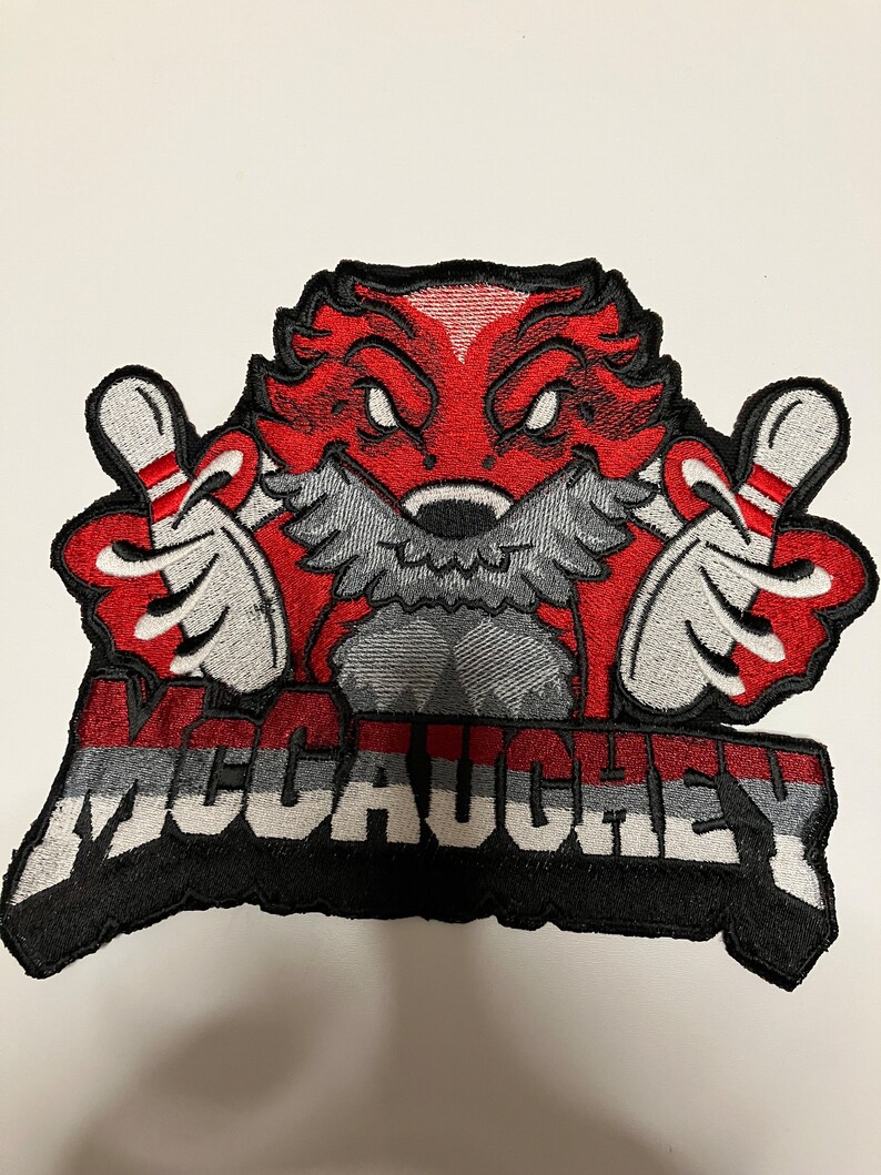 K&ouml;nnte beinhalten: Roter und schwarzer gestickter Aufn&auml;her mit einer Comicfigur, die zwei Bowling-Pins h&auml;lt. Die Figur hat einen roten K&ouml;rper, einen grauen Bart und einen schwarzen Umriss. Der Text "McCauley" ist in wei&szlig;en Buchstaben auf einem schwarzen und grauen Hintergrund geschrieben.