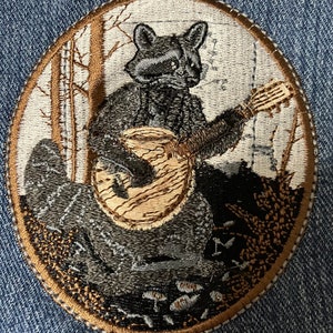 K&ouml;nnte beinhalten: Gestickter Aufn&auml;her mit einem Waschb&auml;ren, der in einer Waldlandschaft Gitarre spielt. Der Waschb&auml;r ist grau und schwarz, die Gitarre ist braun. Der Hintergrund ist eine wei&szlig;e und braune Waldszene.