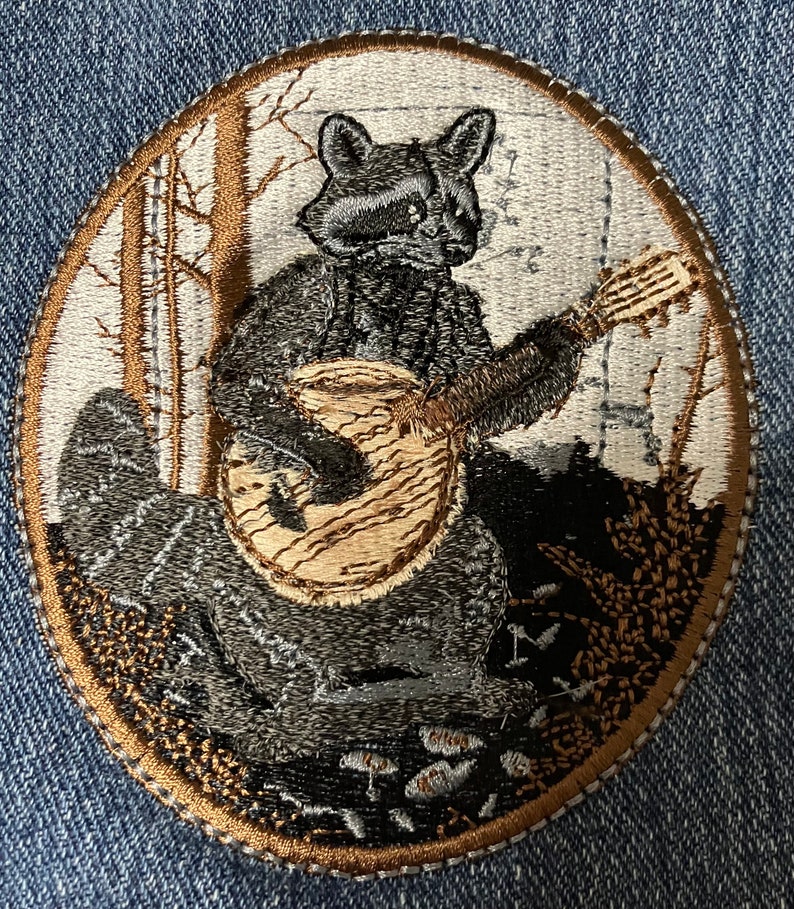 K&ouml;nnte beinhalten: Gestickter Aufn&auml;her mit einem Waschb&auml;ren, der in einer Waldlandschaft Gitarre spielt. Der Waschb&auml;r ist grau und schwarz, die Gitarre ist braun. Der Hintergrund ist eine wei&szlig;e und braune Waldszene.