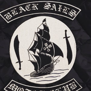K&ouml;nnte beinhalten: Schwarz-wei&szlig; gestickter Patch mit dem Text "BLACK SAILS" und einem Schiff mit einem Kreuz auf seinem Segel. Das Schiff ist von Wellen und zwei Schwertern umgeben.