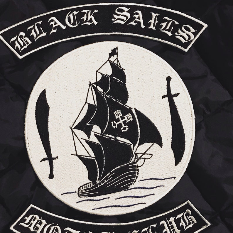 K&ouml;nnte beinhalten: Schwarz-wei&szlig; gestickter Patch mit dem Text "BLACK SAILS" und einem Schiff mit einem Kreuz auf seinem Segel. Das Schiff ist von Wellen und zwei Schwertern umgeben.