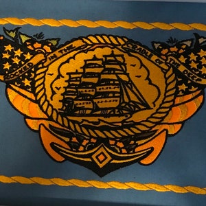 K&ouml;nnte beinhalten: Ein blauer gestickter Aufn&auml;her mit einem goldenen Seilrand. Der Aufn&auml;her zeigt ein Schiff mit Segeln, einen Anker und den Text "ROCKED IN THE CRADLE OF THE DEEP".