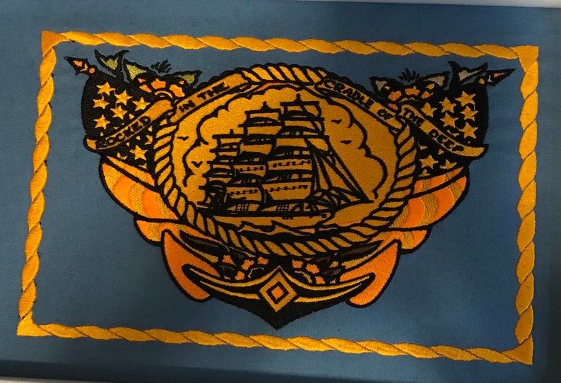 K&ouml;nnte beinhalten: Ein blauer gestickter Aufn&auml;her mit einem goldenen Seilrand. Der Aufn&auml;her zeigt ein Schiff mit Segeln, einen Anker und den Text "ROCKED IN THE CRADLE OF THE DEEP".