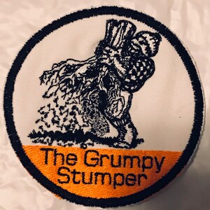K&ouml;nnte beinhalten: Ein wei&szlig;er runder Patch mit schwarzem Rand. Der Patch zeigt eine Schwarzwei&szlig;illustration einer m&uuml;rrisch aussehenden Figur mit einem gro&szlig;en Hut, die vor einem Baum steht. Der untere Teil des Patches hat ein orangefarbenes Band mit dem Text "The Grumpy Stumper" in Schwarz.